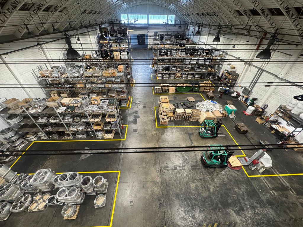 Industrial warehouse with forklifts, metal shelving, and marked floor zones.