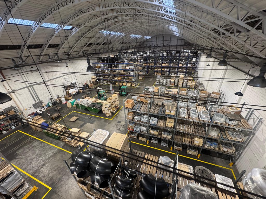 Industrial warehouse with high shelving, technical materials, and marked floor pathways.